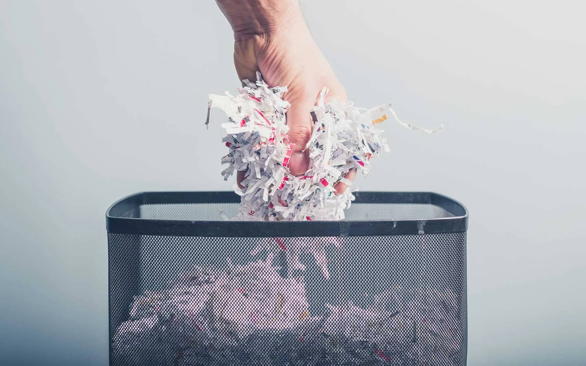 A hand dropping shredded paper into a mesh wastebasket.